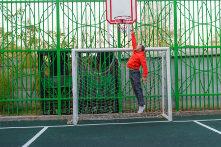 A man in an orange jacket jumps mid-air towards a basketball hoop on an outdoor sports court. He is positioned inside a soccer goal.の写真素材