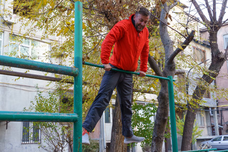 A middle-aged man in an orange jacket and gray pants performs a muscle-up on a green horizontal bar...