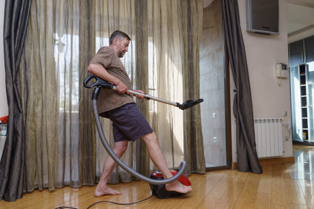 A barefoot middle-aged man playfully uses a vacuum cleaner like a guitar while cleaning the wooden floor in his home.の写真素材