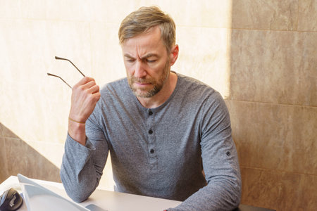 A pensive middle-aged man with a beard, wearing a gray shirt, sits at a desk looking down with a focused expression while holding his glasses.の写真素材