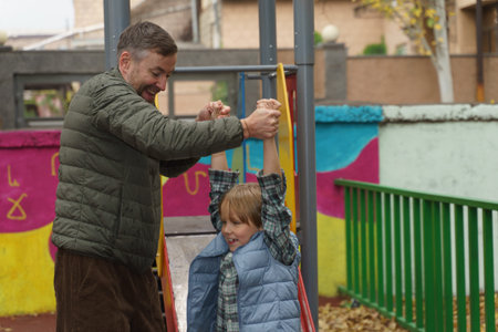 A smiling Caucasian father in a green jacket helps his young son, who is hanging by his hands at the...