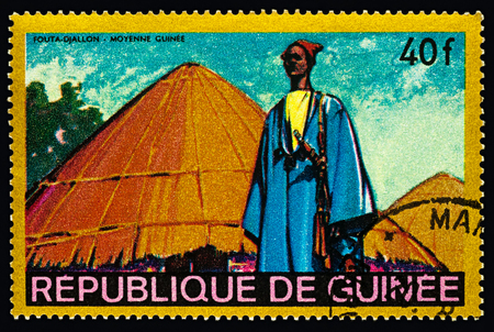 Moscow, Russia - February 27, 2018: A stamp printed in Guinea shows man in traditional clothes in African village, Fouta-Djallon - Middle Guinea, series "Regional Costumes and Habitations", circa 1968のeditorial素材