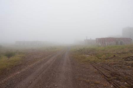 Deserted dirt road through abandoned settlement in tundra, destroyed buildings, pieces of rusty cable, heavy fogの写真素材