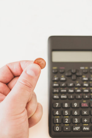 Man's hand holding a one cent euro coin next to a calculator. Financial crisis concept, white...