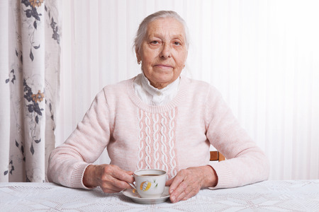 An elderly woman drinks tea at home. Senior woman holding cup of tea in their hands at table closeupの写真素材