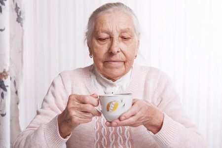 An elderly woman drinks tea at home. Senior woman holding cup of tea in their hands at table closeupの写真素材
