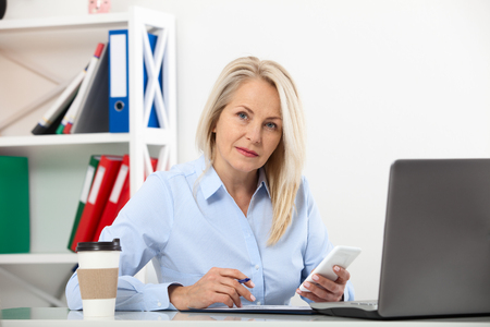 Listening to clients requirements. Beautiful middle aged woman talking on smart phone and smiling at her working place siting in the office.の写真素材