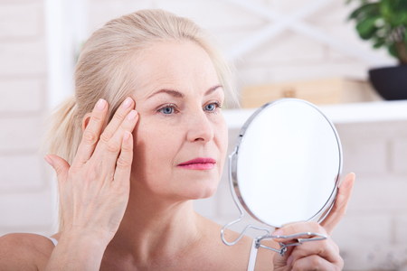 Forty years old woman looking at wrinkles in mirror. Plastic surgery and collagen injections. Makeup. Macro face. Selective focusの写真素材