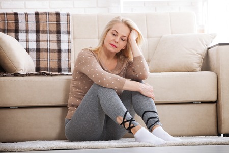 Middle aged barefoot woman sitting at the floor embracing her knees, near sofa at home, her head down, bored, troubled with domestic violence.の写真素材