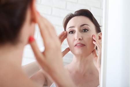 Woman looking at wrinkles in mirror. Plastic surgery and collageの写真素材