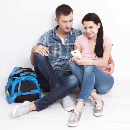 A woman and a man are sitting in a new apartment using a smartphone. A young couple chooses a wedding trip.の写真素材