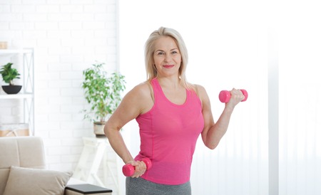 Happy middle aged woman lifting dumbbells at home in the living roomの写真素材