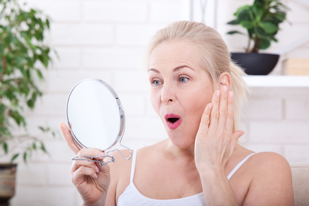 Forty years old woman looking at wrinkles in mirror. Plastic surgery and collagen injections. Makeup. Macro face. Selective focusの写真素材