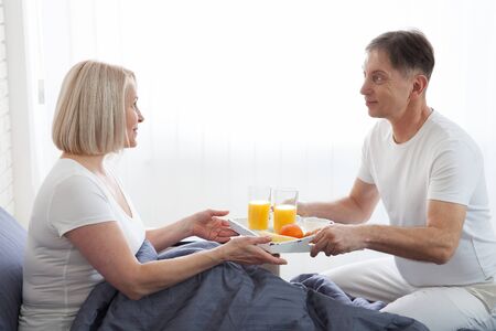 Beautiful couple having breakfast lying in bed at home. Handsome man and attractive woman are enjoying spending time together while gently hugging and kissing sitting in bed.の写真素材