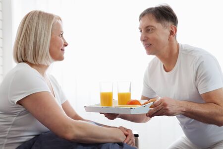 Beautiful couple having breakfast lying in bed at home. Handsome man and attractive woman are enjoying spending time together while gently hugging and kissing sitting in bed.の写真素材
