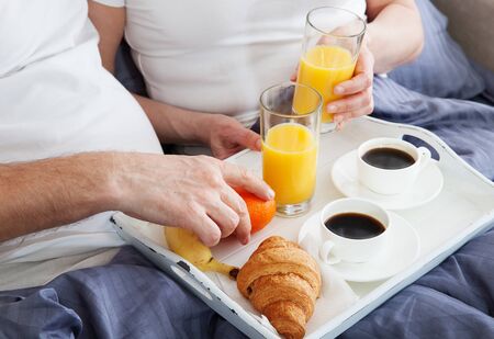 Beautiful couple having breakfast lying in bed at home. Handsome man and attractive woman are enjoying spending time together while gently hugging and kissing sitting in bed.の写真素材