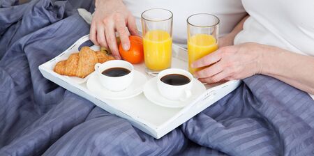 Beautiful couple having breakfast lying in bed at home. Handsome man and attractive woman are enjoying spending time together while gently hugging and kissing sitting in bed.の写真素材