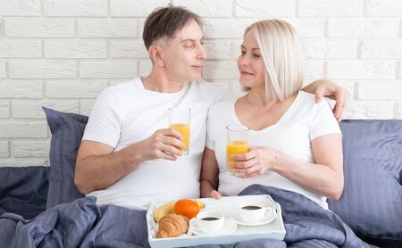 Beautiful couple having breakfast lying in bed at home. Handsome man and attractive woman are enjoying spending time together while gently hugging and kissing sitting in bed.の写真素材