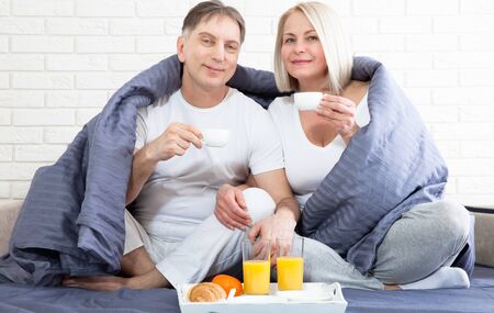 Portrait of happy playful couple relaxing in comfortable cozy bed looking at each other, cheerful man and woman having fun posing. Holding cups of coffee while covering their shoulders with blanket.の写真素材