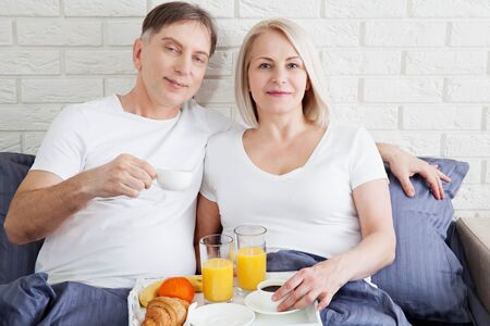 Portrait of happy playful couple relaxing in comfortable cozy bed looking at each other, cheerful man and woman having fun posing. Holding cups of coffee while covering their shoulders with blanket.の写真素材