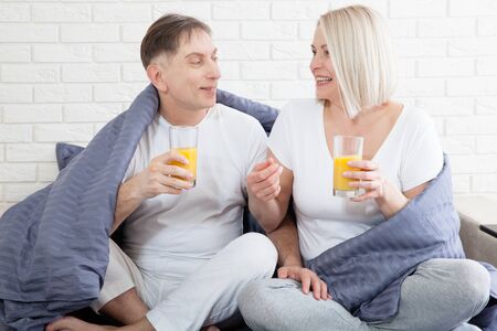 Portrait of happy playful couple relaxing in comfortable cozy bed looking at each other, cheerful man and woman having fun posing. Drink orange juice while covering their shoulders with blanket.の写真素材
