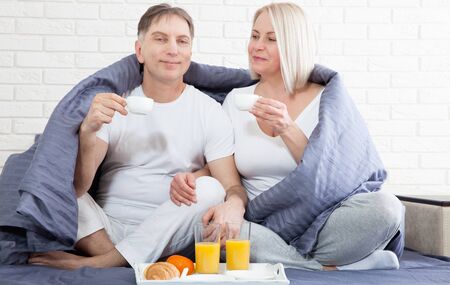 Portrait of happy playful couple relaxing in comfortable cozy bed looking at each other, cheerful man and woman having fun posing. Holding cups of coffee while covering their shoulders with blanket.の写真素材