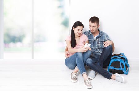 A woman and a man are sitting in a new apartment using a smartphone. A young couple chooses a wedding trip.の写真素材