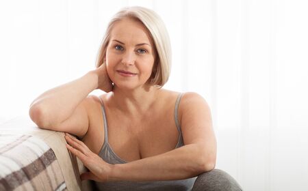 Active beautiful middle-aged woman smiling friendly and looking in camera in living room. Womans face close up. Realistic images without retouching with their own imperfections. Selective focus.の写真素材