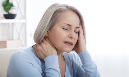 Woman suffering from stress or a headache grimacing in pain as she holds the back of her neck with her other hand to her temple, with copyspace. Concept photo with indicating location of pain.の写真素材