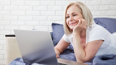 Smiling attractive woman lying on bed using laptop communicating online at home, happy girl typing on computer, enjoying internet shopping or chatting in social networks in bedroom.の写真素材
