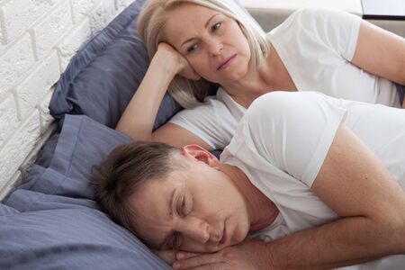 Middle aged woman covering ears while man snoring in bedの写真素材