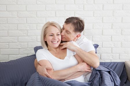 Handsome man and attractive woman are enjoying spending time together while gently hugging and kissing sitting in bed.の写真素材