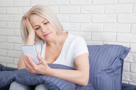 Attractive caucasian woman sitting on bed. Pretty senior using cellphone and resting.の写真素材