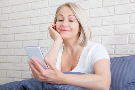 Attractive caucasian woman sitting on bed. Pretty senior using cellphone and resting.の写真素材