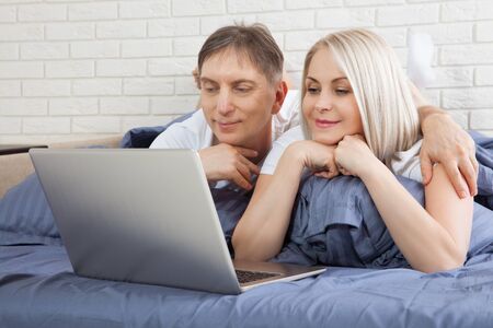 Beautiful amorous couple using laptop, on bed. Beautiful woman and handsome man read the news on internet. Concept relationship.の写真素材