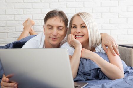 Beautiful amorous couple using laptop, on bed. Beautiful woman and handsome man read the news on internet. Concept relationship.の写真素材