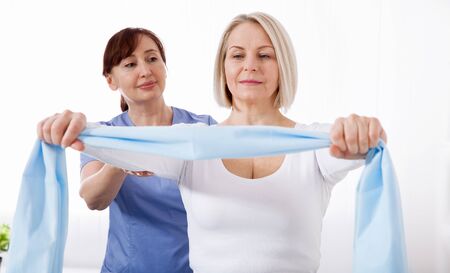 Physiotherapist and woman sitting on a bed exercising with a rubber tapeの写真素材