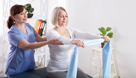 Physiotherapist and woman sitting on a bed exercising with a rubber tapeの写真素材