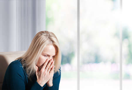 Woman suffering from stress or a headache grimacing in pain. Sad depressed woman at home sitting on the couch, looking down and touching her forehead, loneliness and pain conceptの写真素材