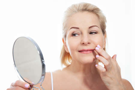 Happy middle aged woman model touching face skin looking in mirror. Smiling mature older lady enjoying healthy skin care, aging beauty, skincare treatment cosmetic products concept.の写真素材