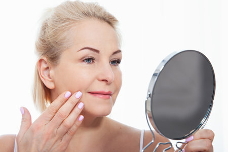 Happy middle aged woman model touching face skin looking in mirror. Smiling mature older lady enjoying healthy skin care. Aging beauty, skincare treatment cosmetic products concept.の写真素材