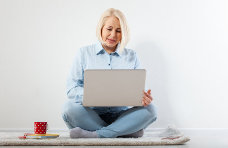 Happy middle aged woman sitting relaxed on the floor using laptop for entertainment. The concept of leisure and work with a cup of coffee at home. front view.の写真素材