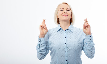 Half-length portrait of beautiful dreamy blonde wearing blue classic blouse standing crossing her fingers with closed eyes wanted to make her wishes come true. Isolated on white.の写真素材