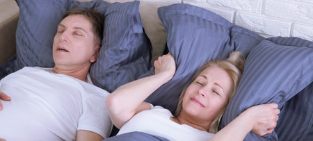 Snoring man. Couple in bed, man snoring and woman can not sleep, covering ears with pillow for snore noise. Middle age couple in bed at home.の写真素材