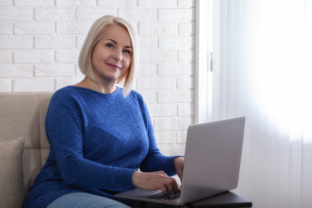 Smiling stylish mature middle aged woman sits with laptop, portrait. Happy older senior businesswoman, looking at camera sitting at home.の写真素材