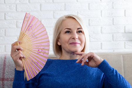 Woman waving a fan pleased with the coolness. Woman enjoying air flow from fan in living room. Mature woman experiencing hot flush from menopause at homeの写真素材