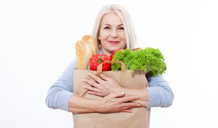 Beautiful middle-aged woman with blond hair holds products from the store in paper bag. Bread, herbs, pepper, green lettuce in her hands for a healthy diet with vitaminswith. Concept healthy foodの写真素材