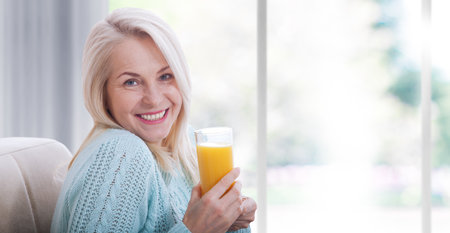 Happy woman holds a glass of orange juice and looks relaxed at the camera, sitting near the window.の写真素材