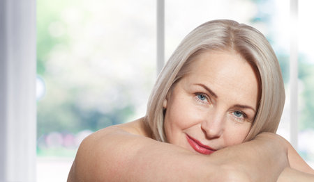 Happy beautiful middle-aged blonde woman shows off her perfectly well-groomed face and sitting relaxed near the window. Realistic images.の写真素材