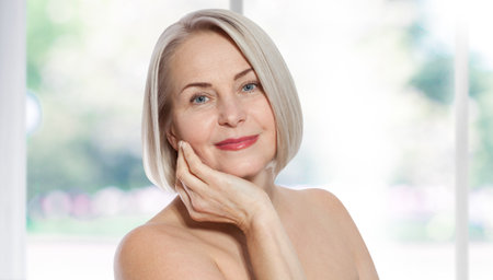 Happy beautiful middle-aged blonde woman shows off her perfectly well-groomed face and sitting relaxed near the window. Realistic images.の写真素材